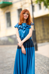 elegant redhead woman in long blue dress posing on old street