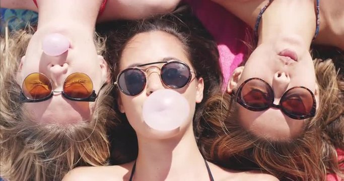 Top view of three teeanage girl friends lying on back blowing bubblegum candy bubbles on beach