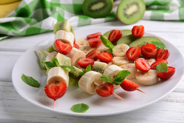 Fresh fruits on skewers in plate on wooden table, closeup