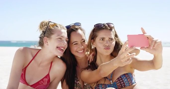 Three teenage girl friends taking selfie on beach making crazy face wearing colorful bikini sharing vacation photo