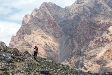 Mountain View and Hiker.
Person with Backpacks Walking Up on Rocky Trail along Green Meadow and High Rock Walls and Summits on Background Blue Sky Clouds Sunny