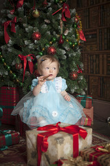Маленькая девочка рядом с новогодней елкой сидит на коробках с подарками 