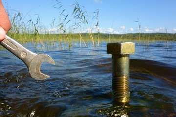 A wrench approaching a sizable metal nut sticking out of a water surface in typical Northern European freshwater lake with water conservation, environmental conservation and ecological concept ideas.