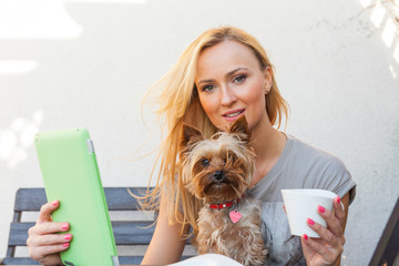 Sensual happy blonde woman sitting on wooden bench with her dog. She is using tablet pc and drinking coffee.