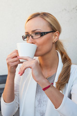Blond hair girl in glasses and suit  sitting on the bench with cup of coffee.