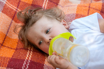 curly child is lying on the coverlet and sucks a bottle of water