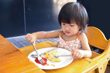 A Little asian girl eating omlet and siting on wooden chair - obrazy, fototapety, plakaty