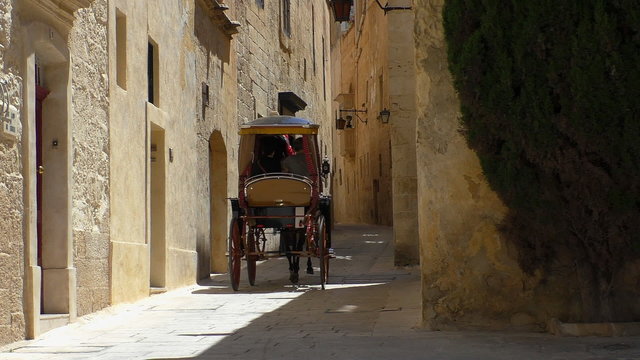 Rear view of one horse drawn carriage with two passengers and the driver wearing straw hat moving slowly away from camera on a narrow street in an old city center centre on a bright sunny day.
