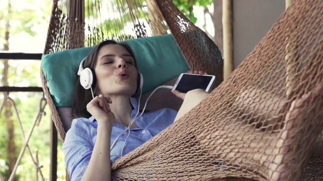 Beautiful, young woman listen to music lying on hammock on terrace
