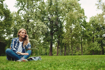 Teenage girl in plaid shirt and denim jeans with smartphone and in park on sunny day talking on the phone laughing.