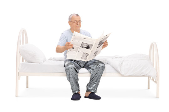 Senior in pajamas reading a newspaper seated on bed