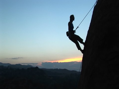 Long-shot of a rock climber silhouetted against a golden-hour-sky rappelling down a cliff.