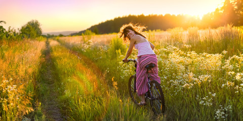 Young woman is riding a bicycle across the sunny field full of flowers. - 91711010