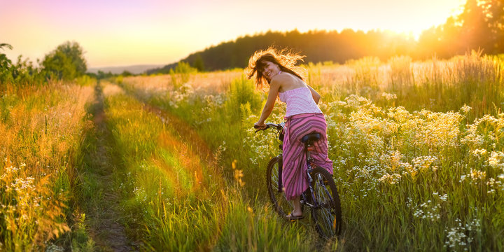 Young woman is riding a bicycle across the sunny field full of flowers.