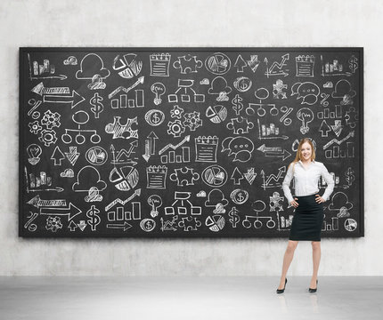 Full length of beautiful young woman with her hands on the waist in formal clothes and a big black chalkboard with drawn business icons behind her. Concrete room.