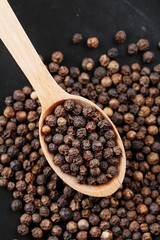 Closeup black pepper whole in wooden spoon on black stone table, top view