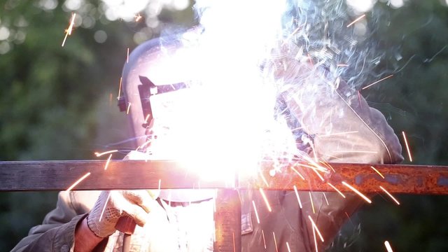 Worker in protective mask welding steel railings outdoors, sparks fly down 
