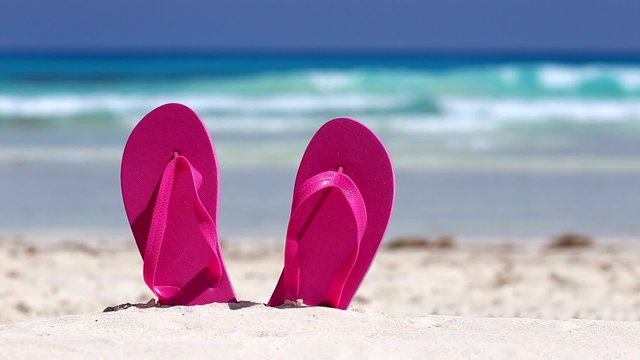 Pink flip flops on white sandy beach near sea waves, nobody. Summer vacation concept
