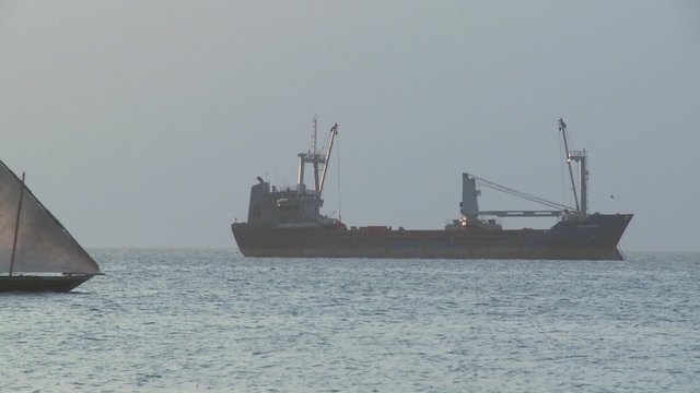 A dhow sailboat sails past a modern freighter cargo ship off the coast of Zanzibar.