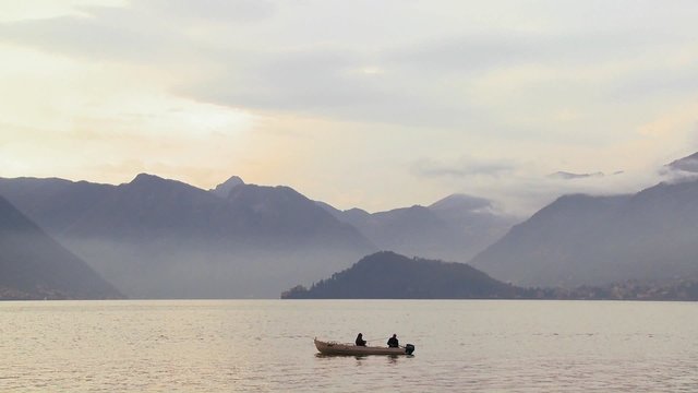Two fishermen fish from a boat on a beautiful lake in front of the Italian Alps.