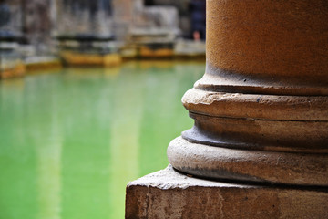 Architectural detail of a stone pillar with a pool of water in the background. Image shot the roman baths, in Bath, England.