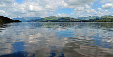 Lake Windermere, seen from a boat. A reflection of the clouds and mountains can be seen on the water. Boats are observable in the water in the far.