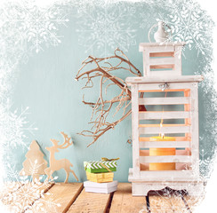 white wooden vintage lantern with burning candle, wooden deer, christmas gifts and tree branches on wooden table. retro filtered image with snowflakes overlay

