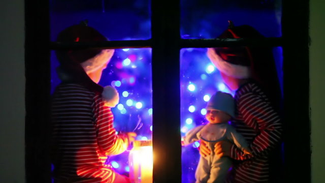 Two children, boys, sitting on a window at night, one holding toy, other playing on phone, waiting for Christmas, colorful tree behind them