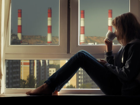 Девушка смотрит в окно на трубы завода и пьет чай или кофе