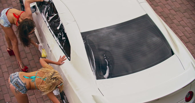High angle view of two attractive young women in bikini washing a white car with sponges 