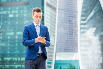 Handsome businessman in suit standing at the street with smart phone in hand. Modern skyscrapers on the background.
