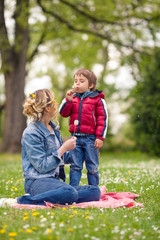 Fototapeta premium Cute little boy blowing dandelion while having a picnic in a park with his mother