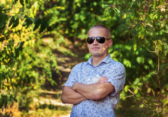 Naklejka na ściany i meble Portrait of handsome man in sunglasses standing in the park with his hands folded
