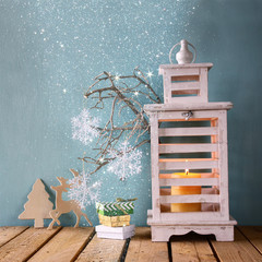 white wooden vintage lantern with burning candle, wooden deer, christmas gifts and tree branches on wooden table. retro filtered image with glitter overlay
