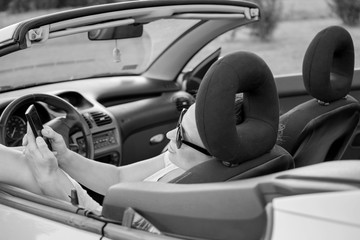 Beautiful young woman in white shirt and black glasses, using mobile phone in a convertible car. Black and white photo. Focus on hands and mobile phone.