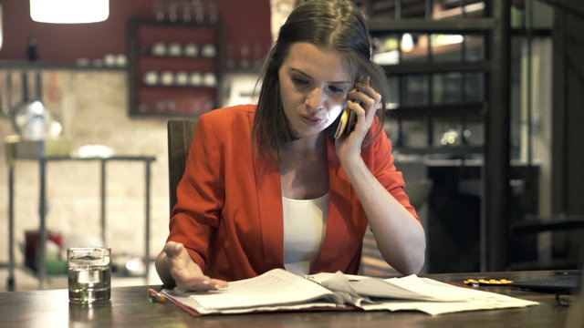 Angry businesswoman with documents talking on cellphone in kitchen at night
