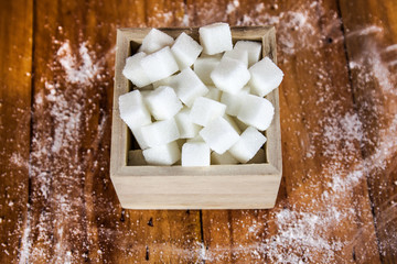 Sugar Cubes in Square Shaped Bowl with Unrefined Sugar spill over in Wooden Background