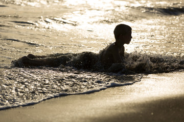 Young boy lays in water at the sandy beach, sunset