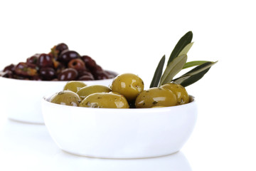 Green and black olives in a white ceramic bowl, with an olive branch and over a white background 