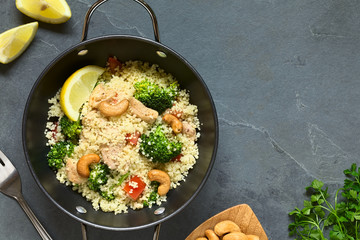 Couscous dish with chicken, broccoli, tomato, cashew nuts. Lemon, cashew nuts and parsley on the side, photographed overhead on slate with natural light.