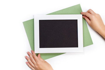 hands holding horizontal white photo frame with black field and green paper under angle on white background isolated with real shadows