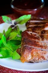 Grilled beef steak with salad and sauce on wooden table at white plate. Salad with tomatoes and cucumbers and sauce barbecue. Leafs of basil as decoration.