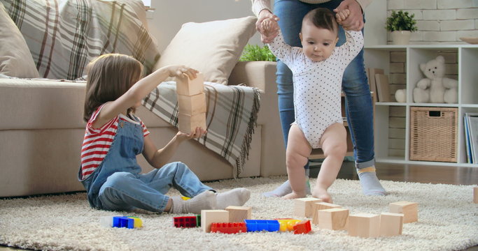 Little girl playing with toy blocks while her baby brother walking helped by mother and then giving him a hug