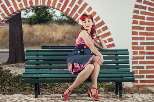 View of pinup young woman in vintage style clothing  sitting on bench on a park.
