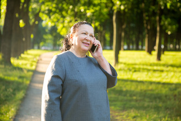 Mature woman using smartphone sitting on a bench in the park. Smiling and talking on the phone