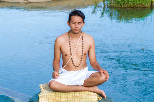 A young brahmin sits in meditation on a river bank in India 
