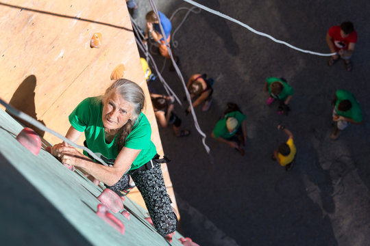 Aged Lady Doing Extreme Sport Elderly Female Moving Up on Outdoor Climbing Wall Sporty Clothing on Fitness Training Intense but Positive Face Using Rope and Belaying Gear