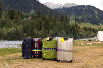 Travel Bags Staying on Wild Grassy Meadow Mountain View with Forest Hills and River Stream and Many Suitcases of different type and colors