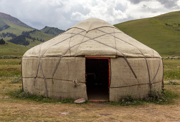 Yurt in Central Asian Veld Traditional Mongol Nomad Housing assembled on Green Meadow among High Mountain Hills in Kyrgyzstan with Opened Entrance door and Yellow Steppe Grass on foreground