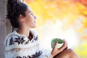 Young beautiful brunette smiling woman with cup of coffee wearing knitted nordic print poncho sitting home by the window. Blurred garden fall background.